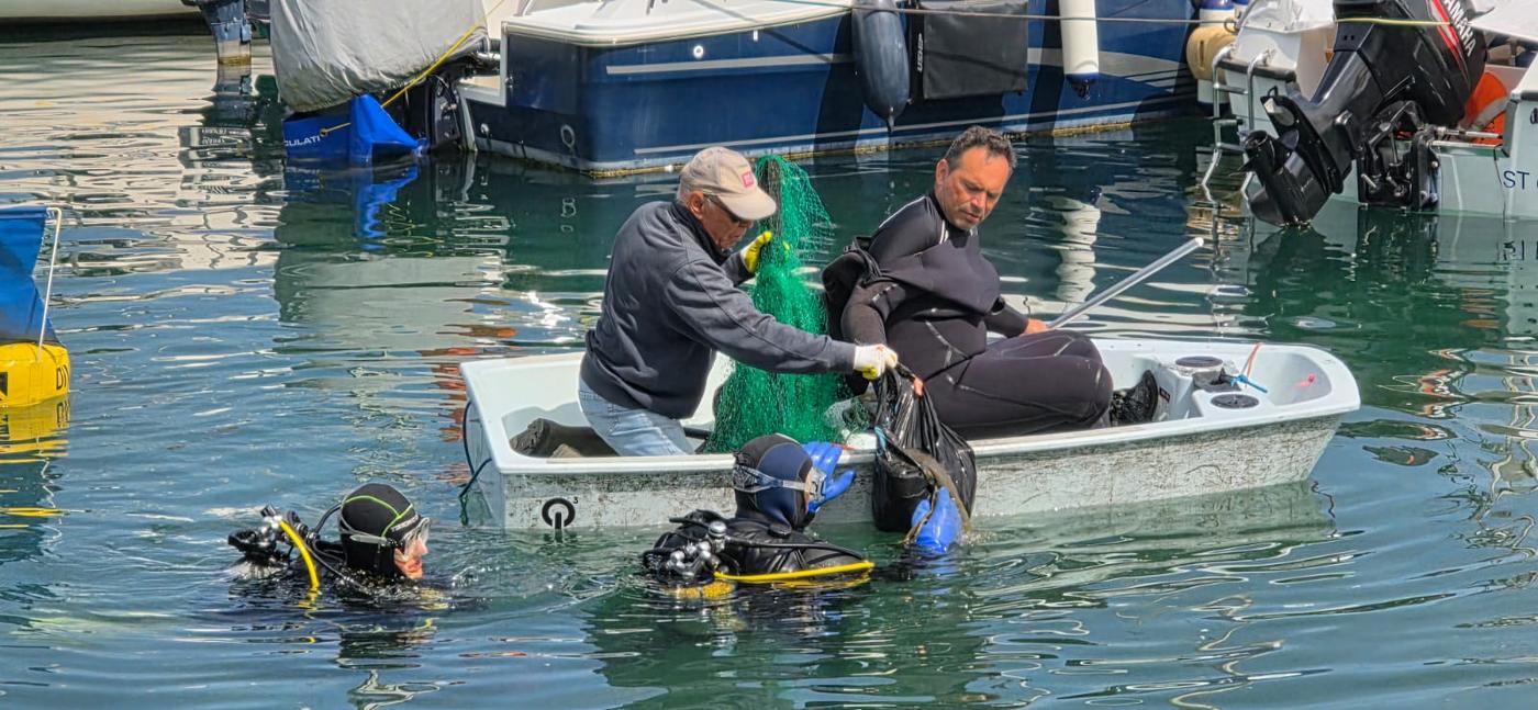 Plongeurs remontant un filet et des déchets du fond du port, aidés depuis une barque